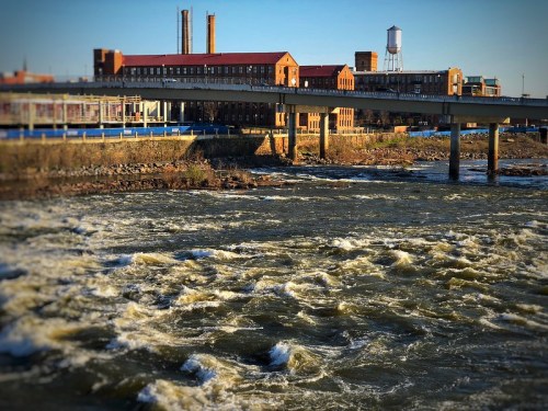 A river with turbulent water flows beneath a bridge, with industrial buildings and a water tower in the background.