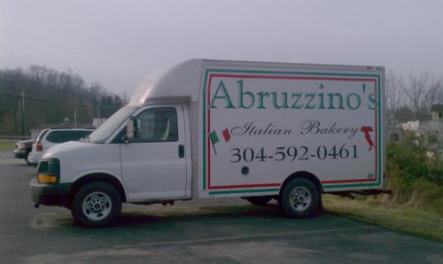 A white delivery truck with "Abruzzino's Italian Bakery" and contact number on the side, parked in a lot.