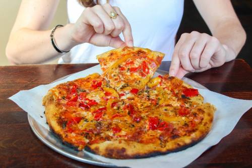 A person pulls a slice of pizza topped with red peppers and cheese from a whole pizza on a wooden table.