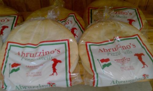 Packages of Abruzzino's pizza shells stacked on a wooden shelf, featuring a red and green logo with an Italian silhouette.