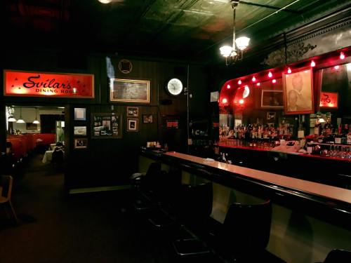 A dimly lit bar area with a long counter, red lights, and vintage decor, featuring a sign for "Svilars Dining Room."