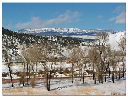 Snow-covered mountains rise in the background, framed by bare trees and a clear blue sky.
