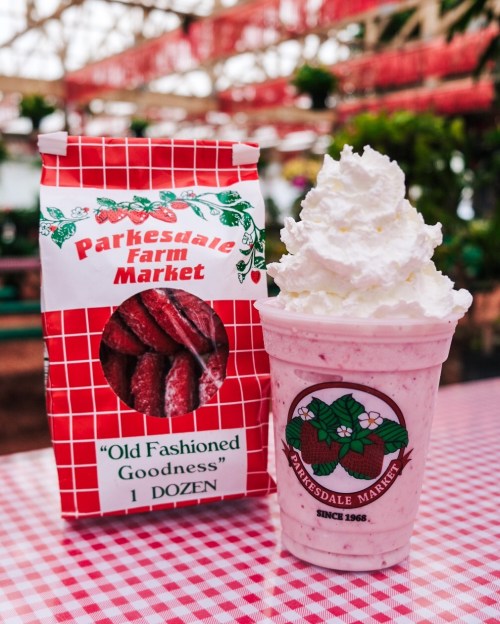 A cup of strawberry milkshake topped with whipped cream next to a bag of strawberry candies on a checkered tablecloth.
