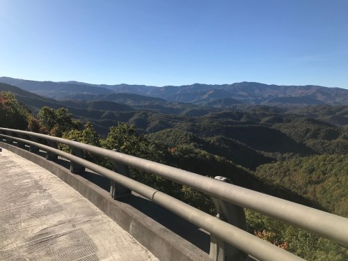 A scenic view of rolling mountains and lush greenery under a clear blue sky, with a curved railing in the foreground.