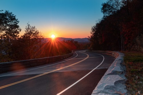 A winding road curves through a landscape at sunset, with vibrant colors and a sunburst in the sky.