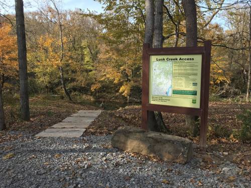 A wooden sign at Lusk Creek Access with a map, surrounded by trees and autumn foliage, near a gravel path.