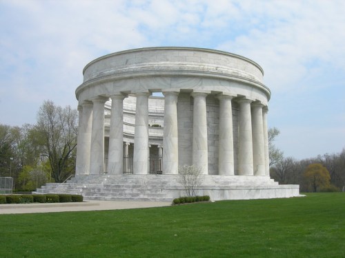 A circular marble monument with columns, set in a grassy area under a cloudy sky.