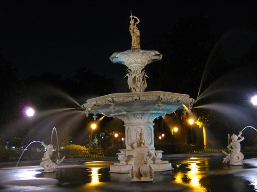 A beautifully illuminated fountain at night, featuring intricate sculptures and water jets against a dark backdrop.
