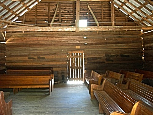 Interior of a rustic wooden building with benches and a door, featuring exposed beams and a simple, natural design.