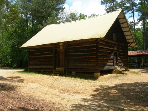 A rustic log cabin with a green metal roof, surrounded by trees and gravel pathways.