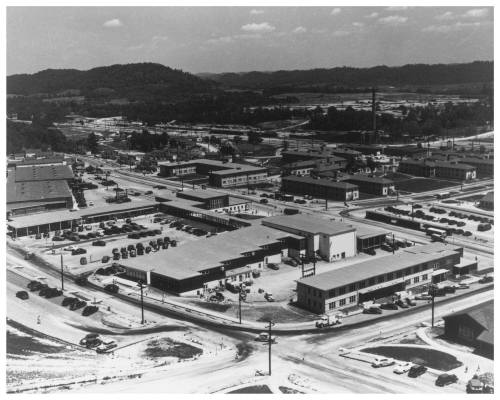 Aerial view of a mid-20th century industrial area with buildings, roads, and parked cars surrounded by hills.