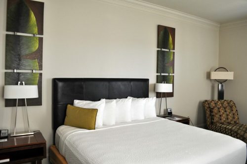 A modern hotel room featuring a bed with white linens, decorative wall art, lamps, and a patterned chair.