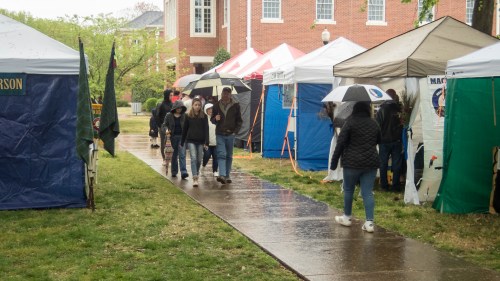 People walking under umbrellas along a wet path lined with colorful tents during a rainy event.