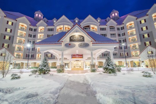A snowy scene of a grand hotel with a peaked roof, warmly lit windows, and festive decorations at dusk.