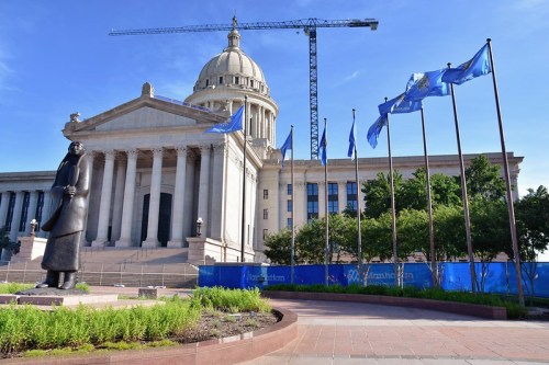 Statue in front of a large government building with flags and construction in the background under a clear blue sky.