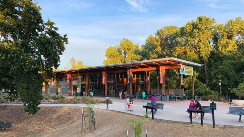 A modern building with large windows surrounded by trees, featuring outdoor seating and people enjoying the space.