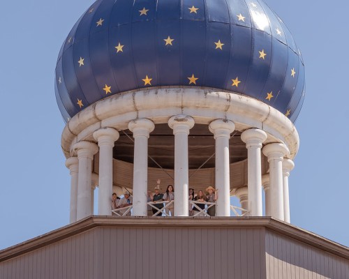 A group of people stands on a balcony beneath a blue dome adorned with stars, supported by white columns.