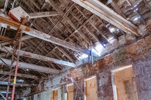Interior of an old, dilapidated building with exposed beams, peeling walls, and light streaming through gaps in the roof.