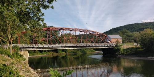 A red arch bridge spans a calm river, surrounded by greenery and hills under a blue sky with wispy clouds.