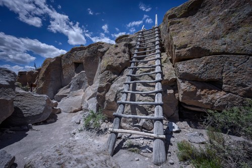 A wooden ladder leads up a rocky terrain under a blue sky with scattered clouds.