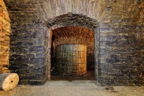 A stone archway leads to a dimly lit room with a large cylindrical tank in the center, surrounded by rough stone walls.