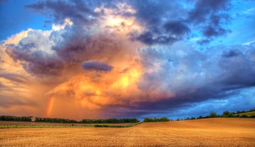A vibrant sunset sky with dramatic clouds and a rainbow over a golden field, creating a serene landscape.