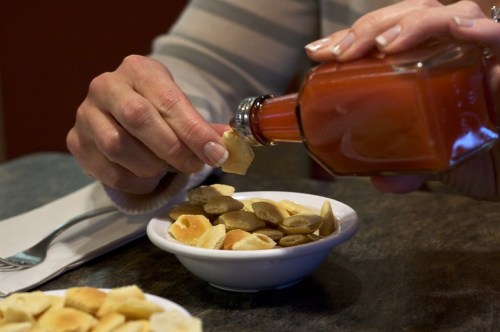 A hand pours sauce over a bowl of assorted snacks on a table.