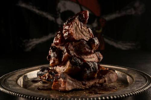 A stack of juicy, grilled meat on a silver platter, set against a dark, pirate-themed background.