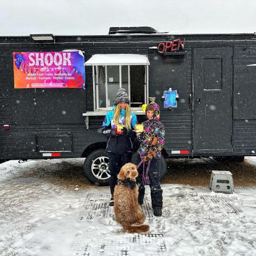 A food truck in the snow with two people and a dog, holding food and wearing winter clothing. Sign reads "SHOOK."