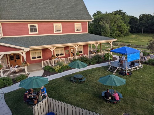 A cozy outdoor gathering with a stage, green umbrellas, and people enjoying a performance near a red building.