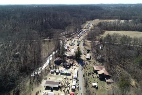 Aerial view of a crowded outdoor event with tents, vehicles parked, and wooded areas in the background.