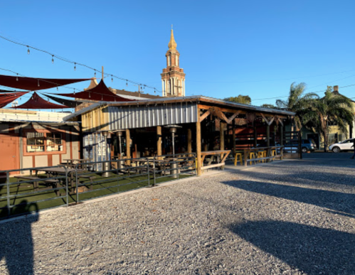 Outdoor dining area with wooden structures, tables, and a tall tower in the background under a clear blue sky.