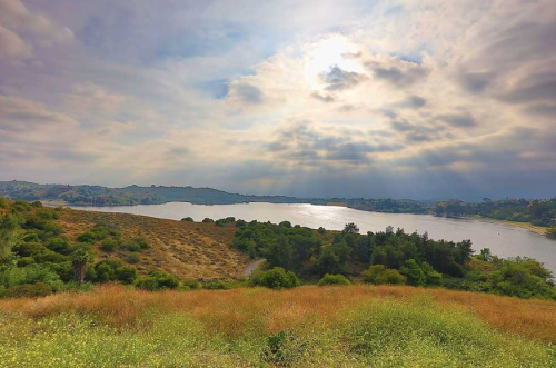 A serene landscape featuring a lake surrounded by hills and lush greenery under a cloudy sky with rays of sunlight.