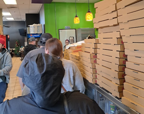 A line of customers waiting at a pizza shop, with stacked pizza boxes visible in the background.