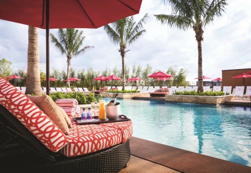 A luxurious poolside scene with lounge chairs, palm trees, and an umbrella, featuring drinks and a champagne bucket.