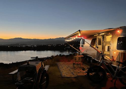 A cozy camper with string lights overlooks a serene lake at sunset, surrounded by mountains.