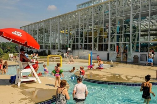 A busy water park scene with children playing in a pool, a lifeguard chair, and a large glass building in the background.