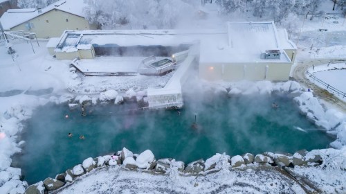Aerial view of a steaming hot spring surrounded by snow-covered landscape and buildings. People are enjoying the water.