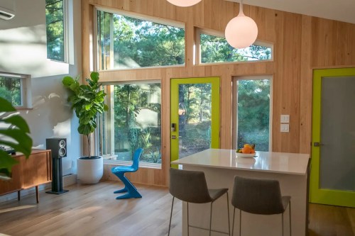 Bright modern kitchen with large windows, green door, and stylish furniture, featuring a plant and fruit on the counter.