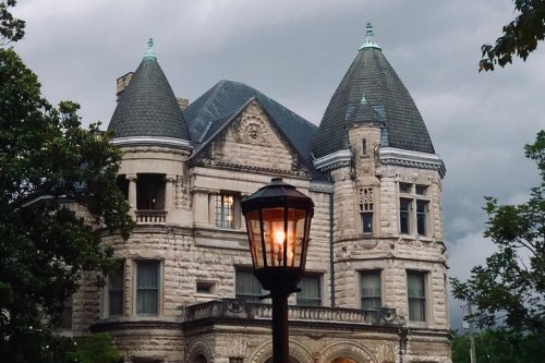 A historic stone mansion with turrets under a cloudy sky, illuminated by a streetlamp in the foreground.