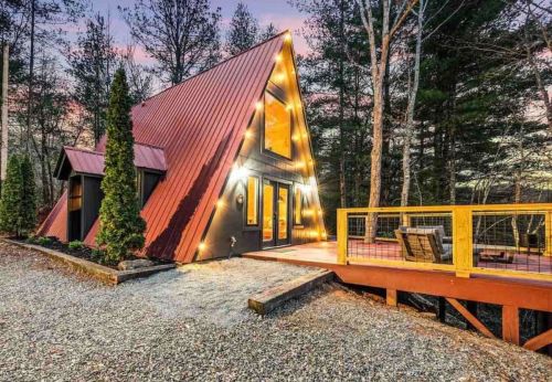A cozy A-frame cabin with a red metal roof, surrounded by trees and featuring a wooden deck with seating.