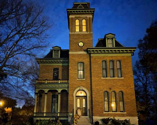 Historic brick building with a tower, illuminated at night, surrounded by trees and a garden.