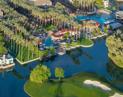 Aerial view of a luxurious resort with palm trees, a pool, and a golf course beside a serene body of water.