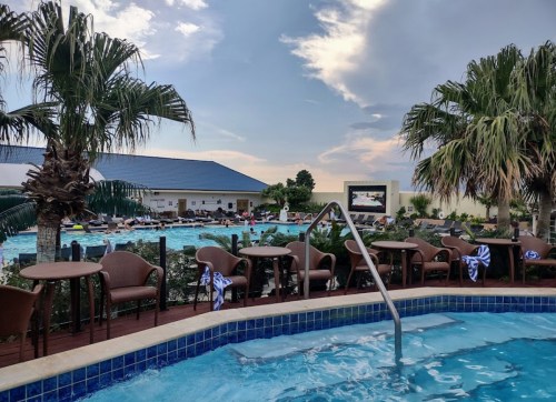 A pool area with palm trees, lounge chairs, and tables, under a cloudy sky. People are enjoying the water.