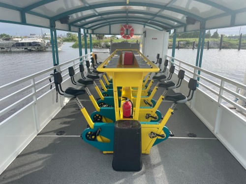Interior of a boat featuring rower seats and a central rowing machine, with a view of water outside.