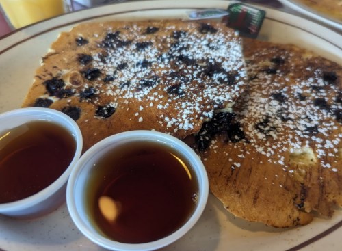 Two large pancakes with blueberries, sprinkled with powdered sugar, served with two small cups of syrup.