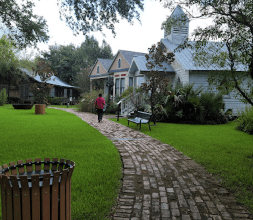 A person walks along a brick path in a lush green garden, surrounded by charming houses and trees.