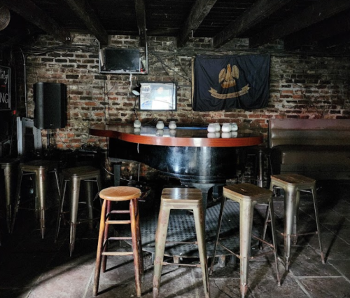 A dimly lit bar with a wooden piano, metal stools, and a brick wall, featuring a flag in the background.