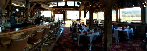 A rustic restaurant interior with wooden beams, a bar, and tables set for dining, featuring large windows and scenic views.