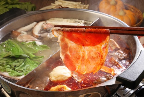 A close-up of a hot pot with various ingredients, including meat and vegetables, being lifted with chopsticks.